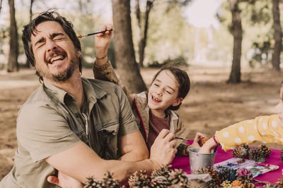 Playful girl coloring father while sitting with pine cones at picnic table in park