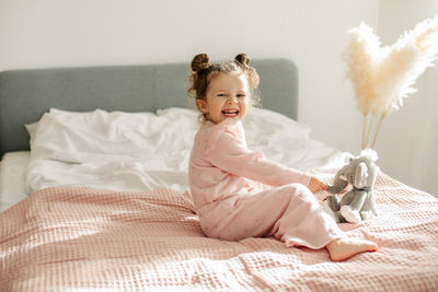 A cheerful little girl is sitting on the bed after waking up and playing with her toy elephant