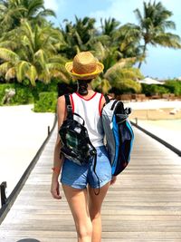 Rear view of woman with backpacks walking on wooden footpath against trees at beach