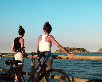 People sitting on bicycle by sea against clear sky