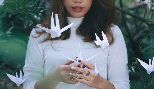 Midsection of young woman hanging flying paper cranes while standing against plants at park