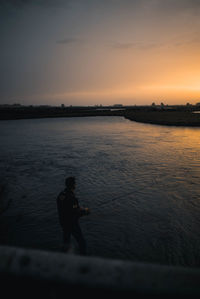 Silhouette man fishing at shore against sky during sunset