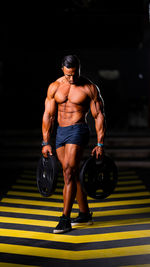 Full length of shirtless man holding weights while walking on floor