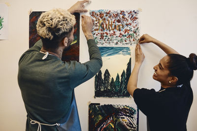 Smiling female and male students sticking painting on wall in art class