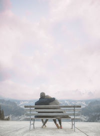 Couple sitting on bench against cloudy sky