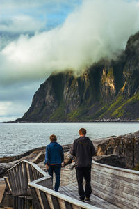 Rear view of friends walking on boardwalk by lake against mountains