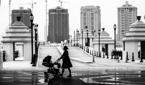 Side view of mother with baby carriage walking against buildings in city