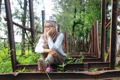 Portrait of young woman sitting on rusty metal against trees