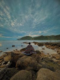 Man sitting on rock at beach against sky during sunset