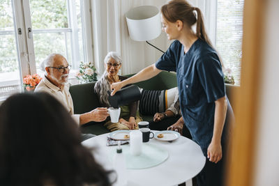 Smiling nurse serving tea to senior man holding mug while sitting in nursing home
