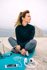 Full length of woman sitting on exercise mat by sea on walkway