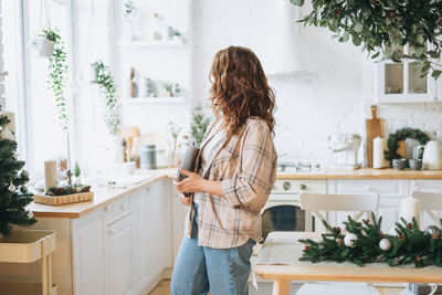 Attractive smiling woman with curly hair in plaid shirt with laptop at bright kitchen at home