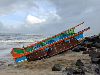 Broken capsized fishing boat moored on beach against sky