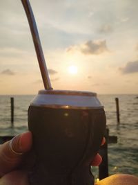 Close-up of hand holding drink at beach against sky during sunset