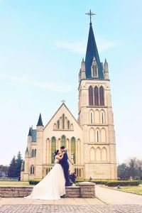 Newly wed couple embracing by church against sky