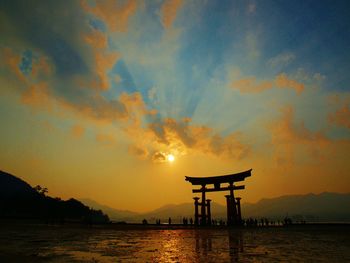 Silhouette torii gate at itsukushima shrine by lake against sky during sunset
