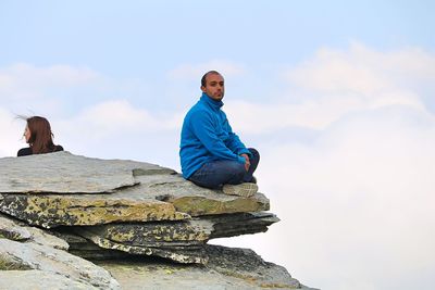 Young man sitting on rock against sky