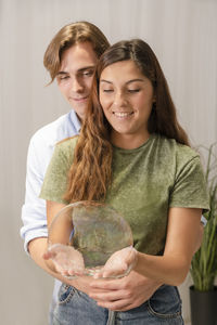 Smiling young couple holding bubble while standing against wall at home