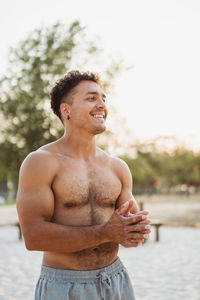 Smiling muscular hispanic ethnic male athlete with naked torso rubbing sand on hands during training on the beach looking away