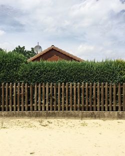 Cottage and plants by fence against cloudy sky