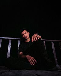 Portrait of young man sitting on railing against black background