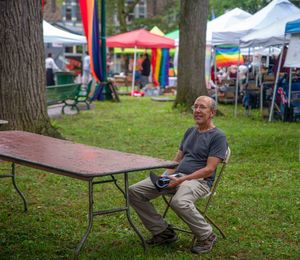 A smiling man sits alone at outdoor pride fest with rainbow flags