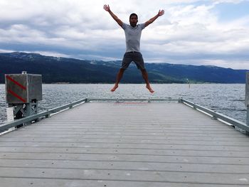 Full length of man jumping with arms outstretched over pier against cloudy sky