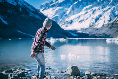 Man skipping rocks.man standing on rock by lake against snowcapped mountains during winter. 