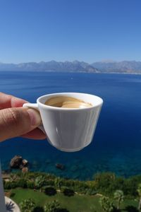 Close-up of hand holding coffee cup by sea against blue sky