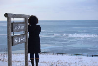 Rear view of woman looking at sea while standing on snowy field against sky