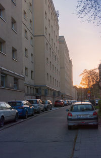Cars on road by buildings against sky during sunset