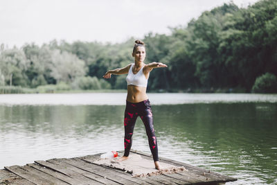 Woman practicing warrior 2 pose while standing on pier over lake in forest