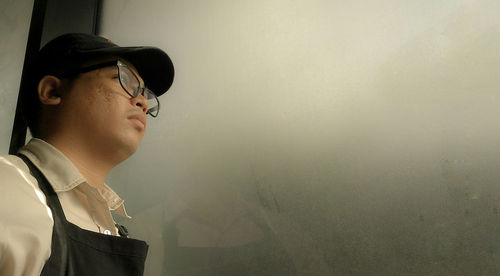 Young man looking away while standing against window