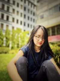 Portrait of asian woman in eyeglasses sitting on the grass with arms hugging legs against buildings.