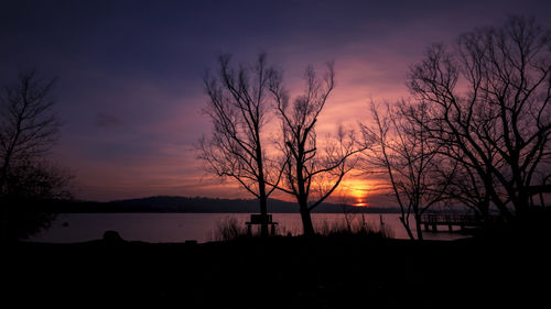 Silhouette bare trees by lake against romantic sky at sunset