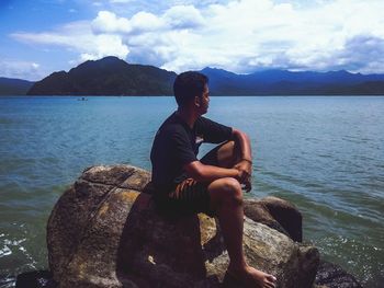 Man sitting on rock looking at sea against sky
