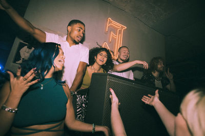Cheerful young multiracial men and women dancing around dj at illuminated nightclub