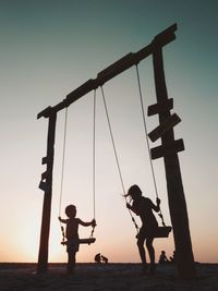 Children swinging on field against sky during sunset