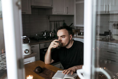 Young man drinking tea and reading a book in his modern light kitchen