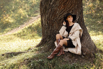 Smiling woman wearing hat sitting by tree at park