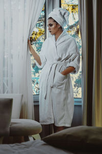 Woman in bathrobe standing by window at home
