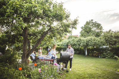 Male and female friends preparing food on barbecue while family having fun in backyard