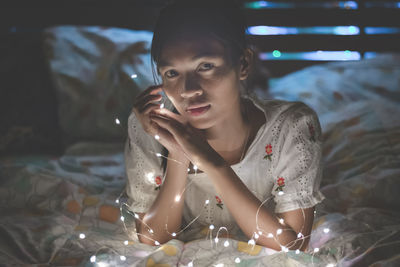 Close-up portrait of young woman lying by illuminated string lights on bed