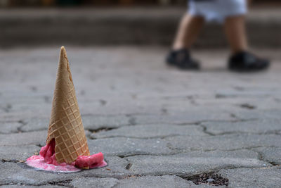 Fallen pink strawberry sorbet ice cream with cracking cone on cement ground and blurry children legs