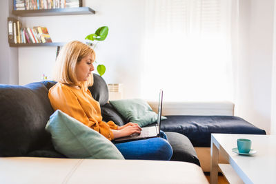Side shot of adult woman in jeans and blouse sitting on sofa in living room browsing netbook