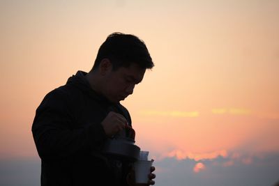 Young man pouring drink in glass against sky during sunset