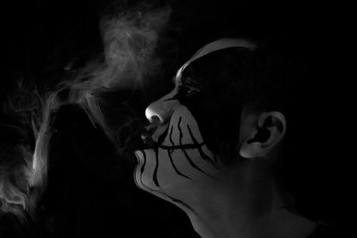 Side view of man with face paint emitting smoke against black background