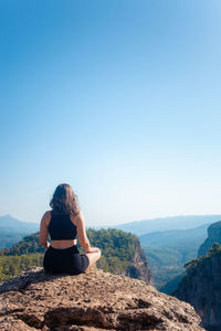 Rear view of woman sitting on rock against clear blue sky