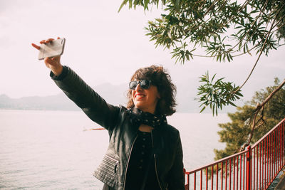 Smiling woman taking selfie with smart phone while standing by lake at park