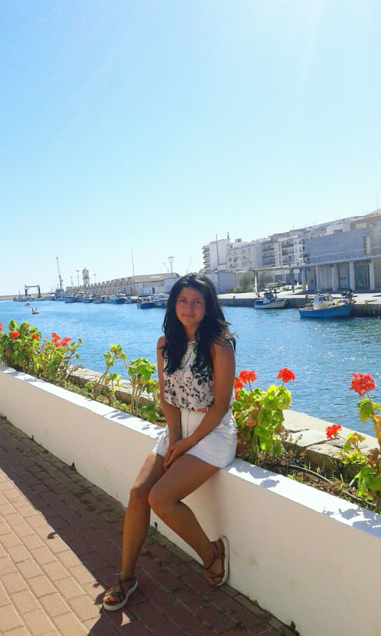 water, clear sky, building exterior, lifestyles, leisure activity, architecture, full length, copy space, young adult, built structure, young women, city, casual clothing, person, portrait, looking at camera, river, day, sunny, weekend activities, sea, in front of, long hair, vacations, outdoors, city life